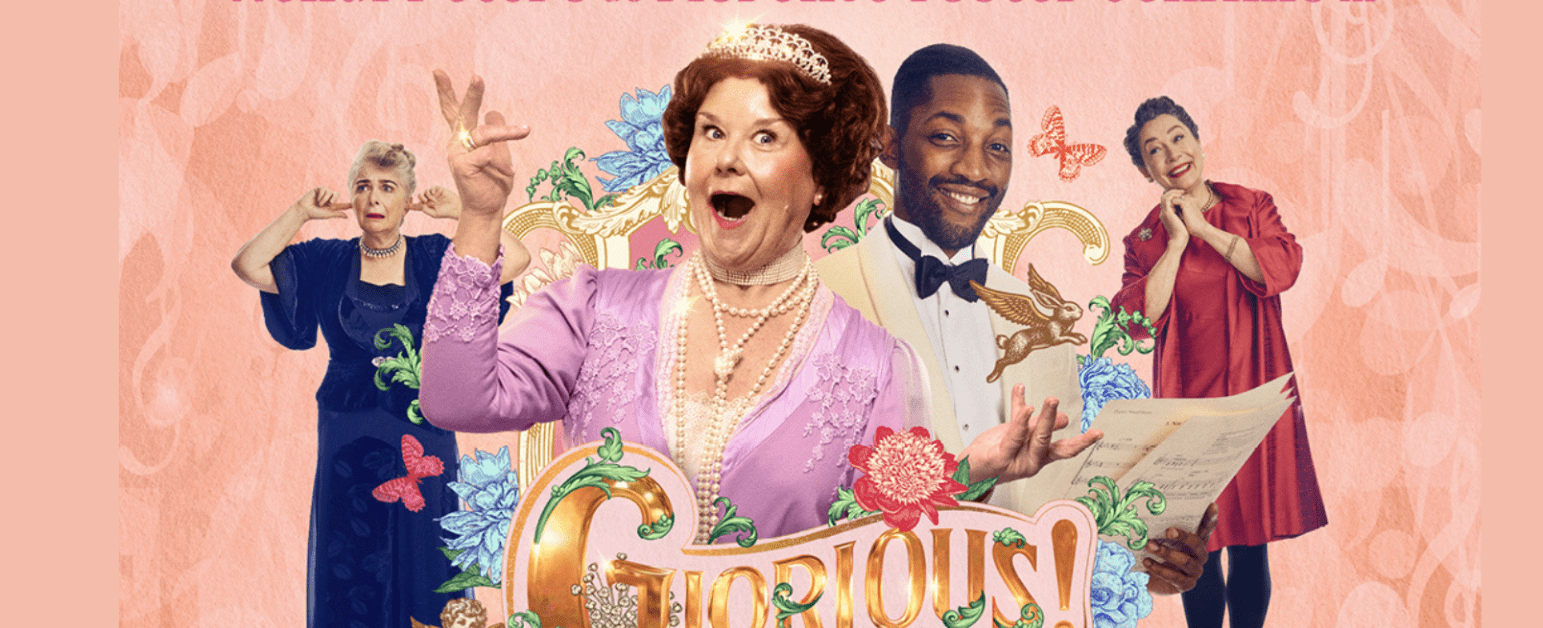 A woman with brown hair and a tiara wearing a pearl necklace and a pink top is singing in front of a pink background with an ornate golden frame, with a smiling man and woman next to her and one woman with her fingers in her ears