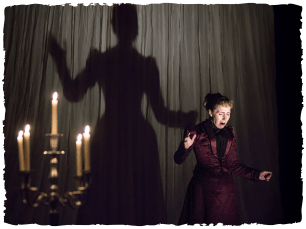 A woman in a black victorian outfit has her shadow projected onto a curtain behind her while stood next to a few candles