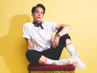 Suzi Ruffell in a white top and black trousers sits on a stool in front of a yellow background