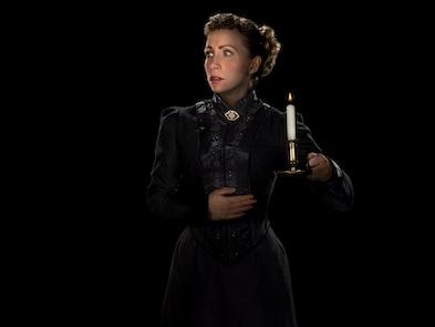 A performer in black victorian dress holding a candelight looking distressed