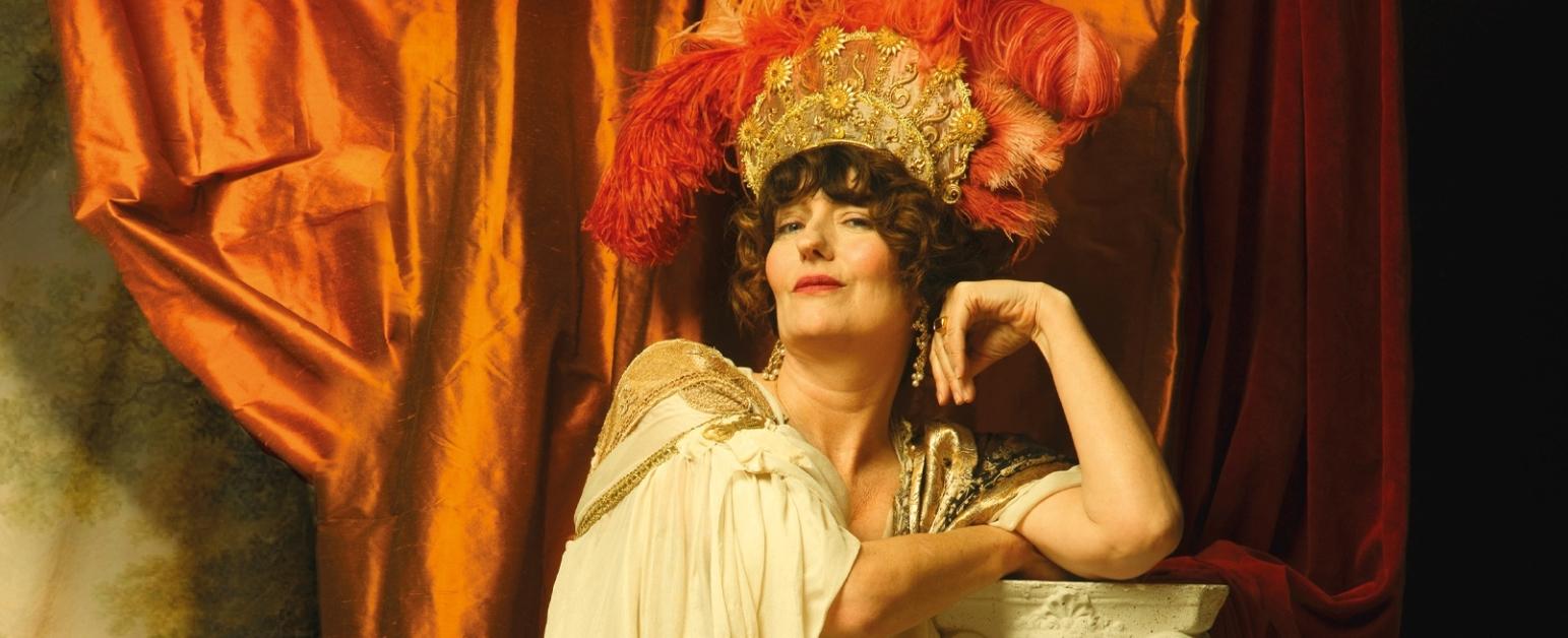 Anna Chancellor with a golden crown with red plumes  and a while dress with gold embroidery leans on a marble column in front of an orange draped curtain