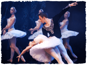 A ballet performer in white being held by another in black as they lean backwards while outstretching their arm