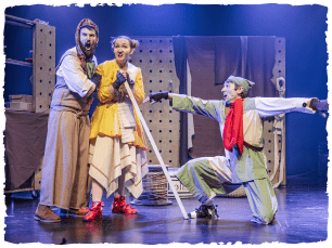 3 people on stage, 2 look scared huddled, one kneeling on floor