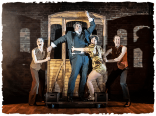 Houdini and characters holding onto the front of  a train onstage