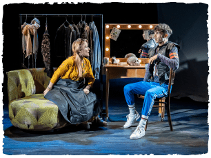 two actors in a dressing room set. one is sitting on a sofa and one at a dressing table