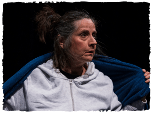 Woman in grey zip up jumper, hair tied up and messy with blue blanket round her, serious expression