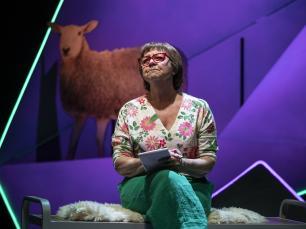 Tessa Peake-Jones sits on a coat, a sheep behind her, and writes a letter to Vera. She wears green trousers and a floral top, looking into the middle distance with a thoughtful expression.