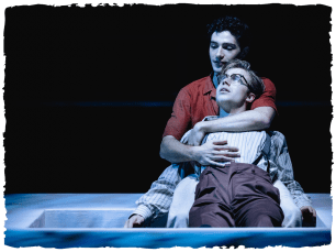 two men sitting on the floor, looking gloomy. One man is leaning into another man's arms.