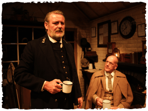 Man stood and man sat down both drinking tea.