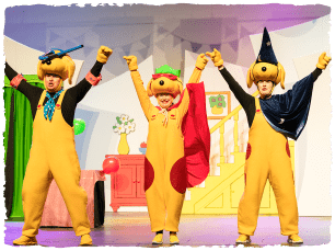 3 actors dressed as dogs. They are also wearing capes and party hats. BEhind them is a set painted as a front room 