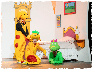 Two actors dressed as dogs. They are sat on the floor at the corner of a bed. Another actor dressed as an alligator is under the bed