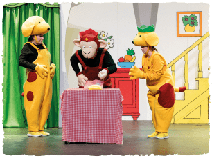 2 actors dressed as dogs. In the middle of them another dressed as a monkey. They are standing round a table with a red checkered tablecloth on it. 