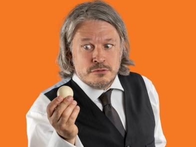 Richard Herring in a suit holding a white snooker ball on an orange background