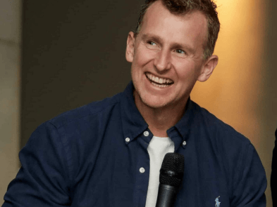 Nigel Owens MBE, a white man with brown hair smiling wearing a blue shirt and a white t-shirt holds a microphone