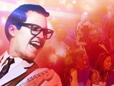 A performer dressed as Buddy Holly with short blakc hair, black rimmed glasses and a white suit holds a guitar in front of a background surrounded by performers