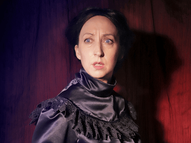 A performer in a black Victorian dress and black hair in front of a dark red background