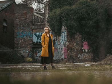 A woman in a yellow coat walks through industrial ruins overgrown with plants