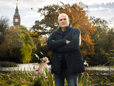 Image of John Crace, with Big Ben in the back and a small blonde dog jumping in the air behind him. Autumnal trees in background.
