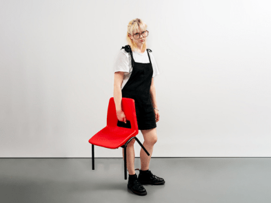 Blonde woman with glasse, in a white tshirt, black doc marten boots and black pinafore, is holding a red school-style chair with a sulky expression. 