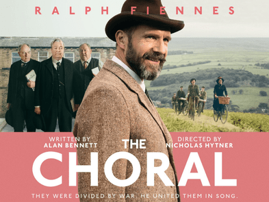 Ralph Fiennes in front of 3 men stood in a line on the left and a field with people cycling on the right