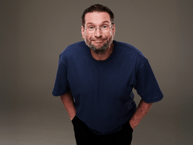 An actor wearing a black t-shirt and glasses leaning towards the camera 