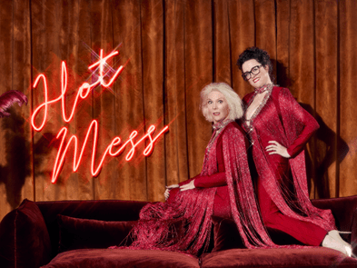 A woman with blonde shoulder length hair, and another with short black hair and glasses. They are wearing red satin clothes and  sitting on the back of a red velvet sofa with a fluorescent sign that says 'hot mess'