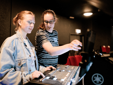 Two women are standing behind a sound desk, one is showing the other how to do something, and they are looking at a screen. 